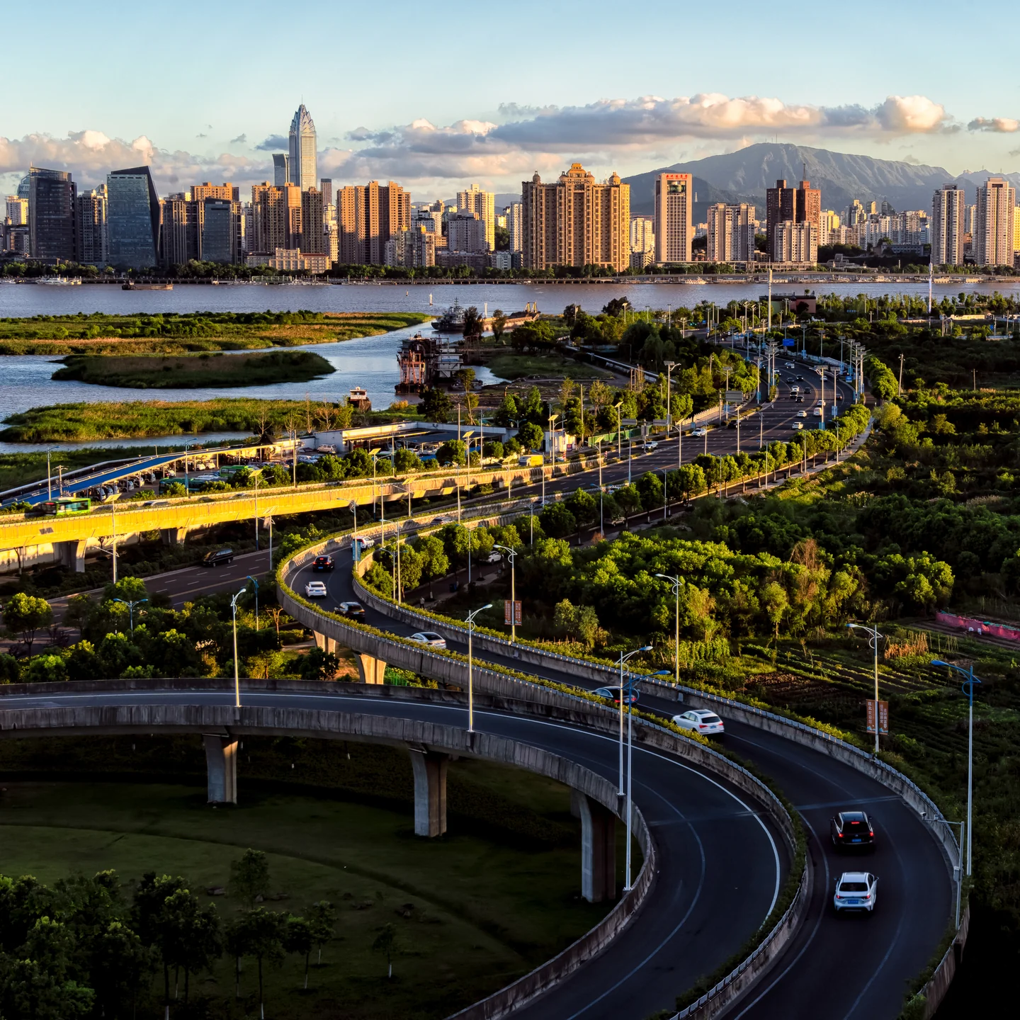 Curving roads lead toward a city skyline beneath sunlit clouds.
