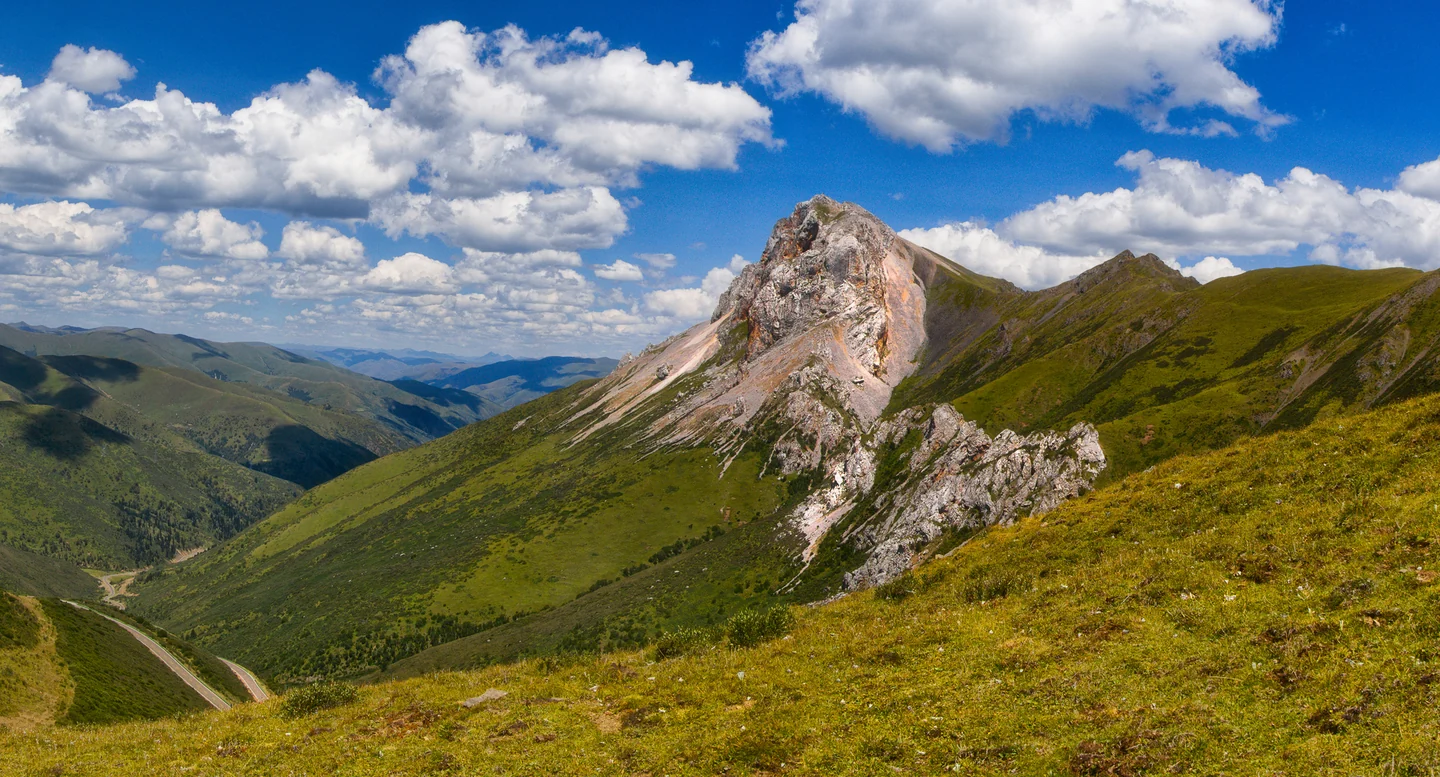 A rocky mountain ridge rises from green grassland beneath blue sky and white clouds.