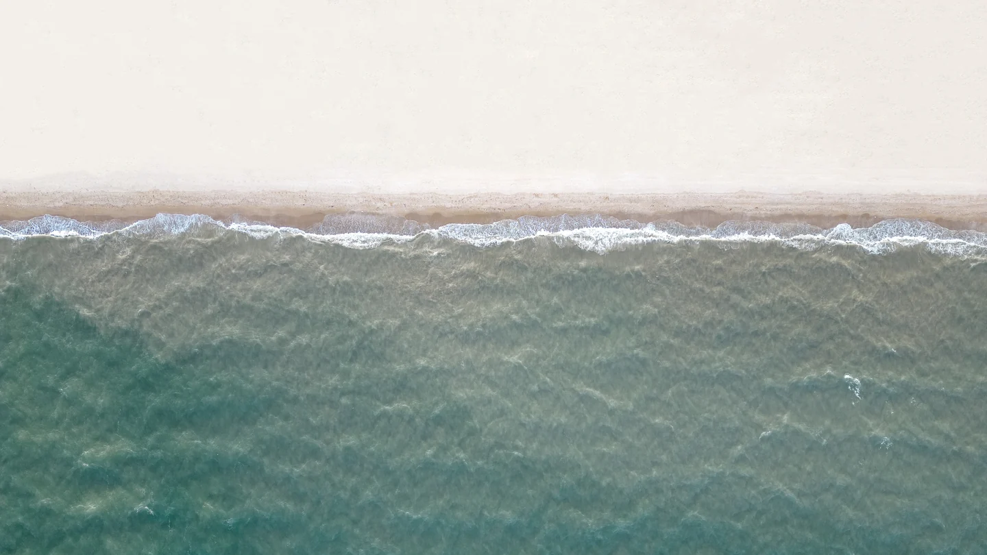 Pale sand and foamy waves meet a textured blue-green sea, viewed from above.