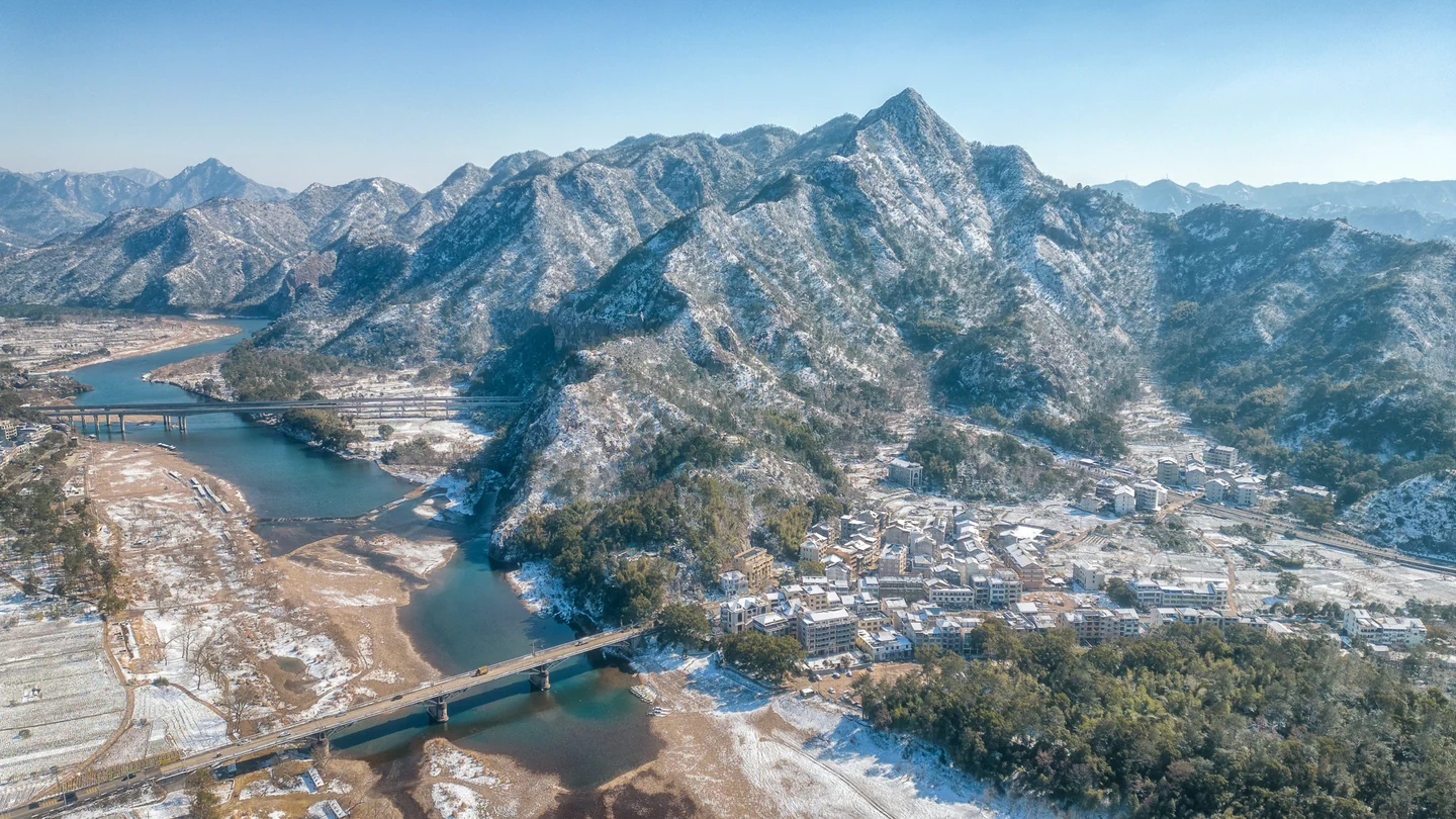 A light blanket of snow covers a village and mountain ridges beneath a blue sky.