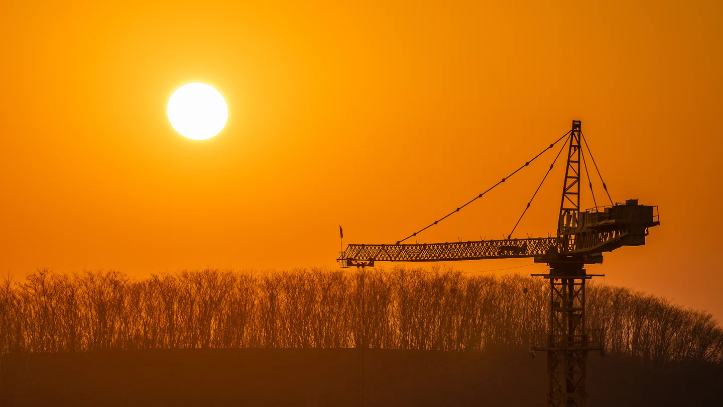 An orange sun rises beside an industrial crane and silhouetted trees.