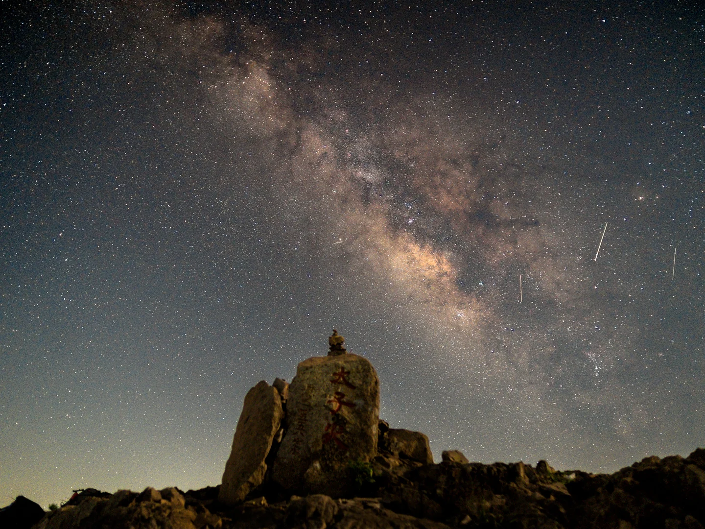 The Milky Way stretches above a rocky summit and a lone figure.
