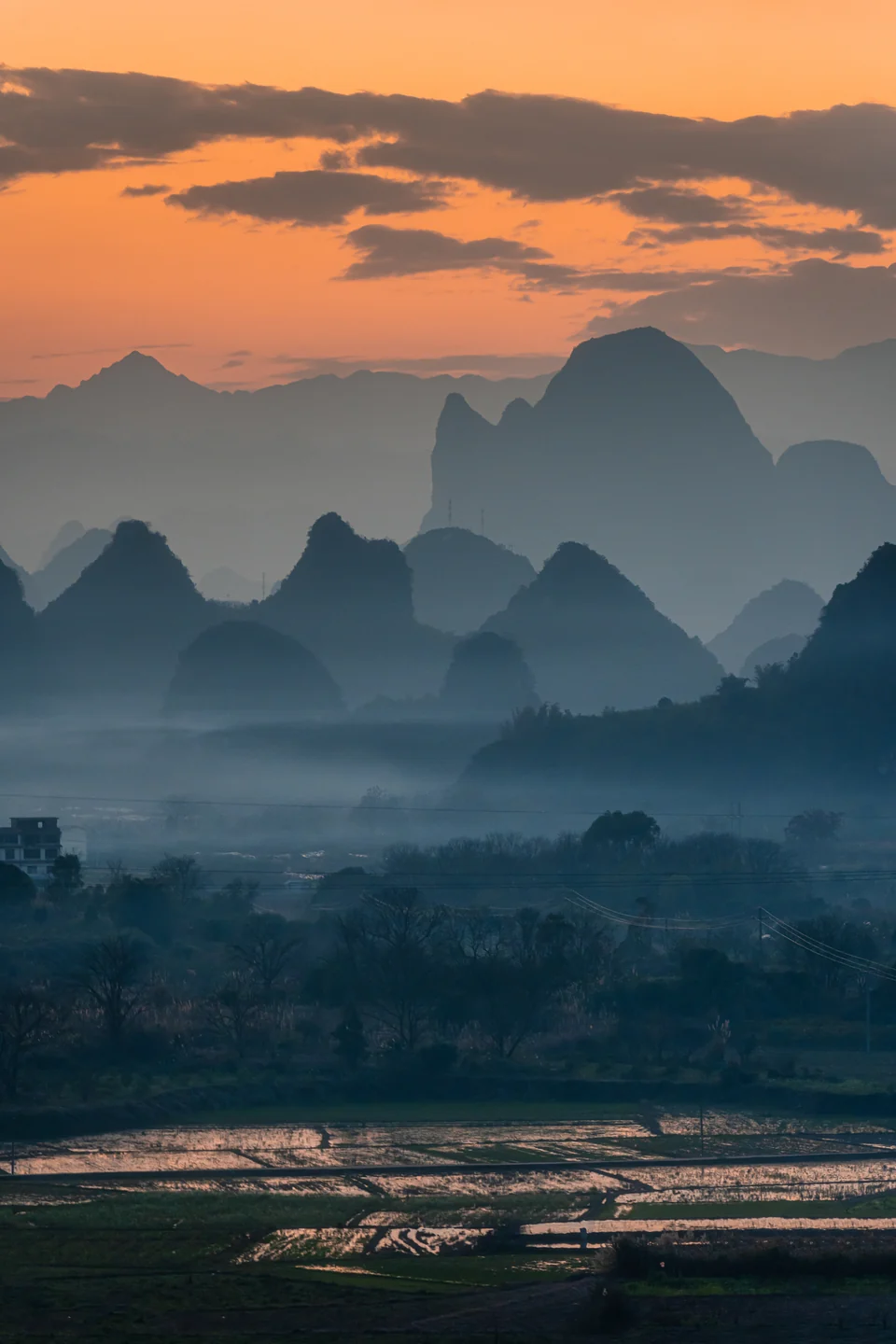 An orange sunset glows over layers of hazy karst mountains and reflective fields.