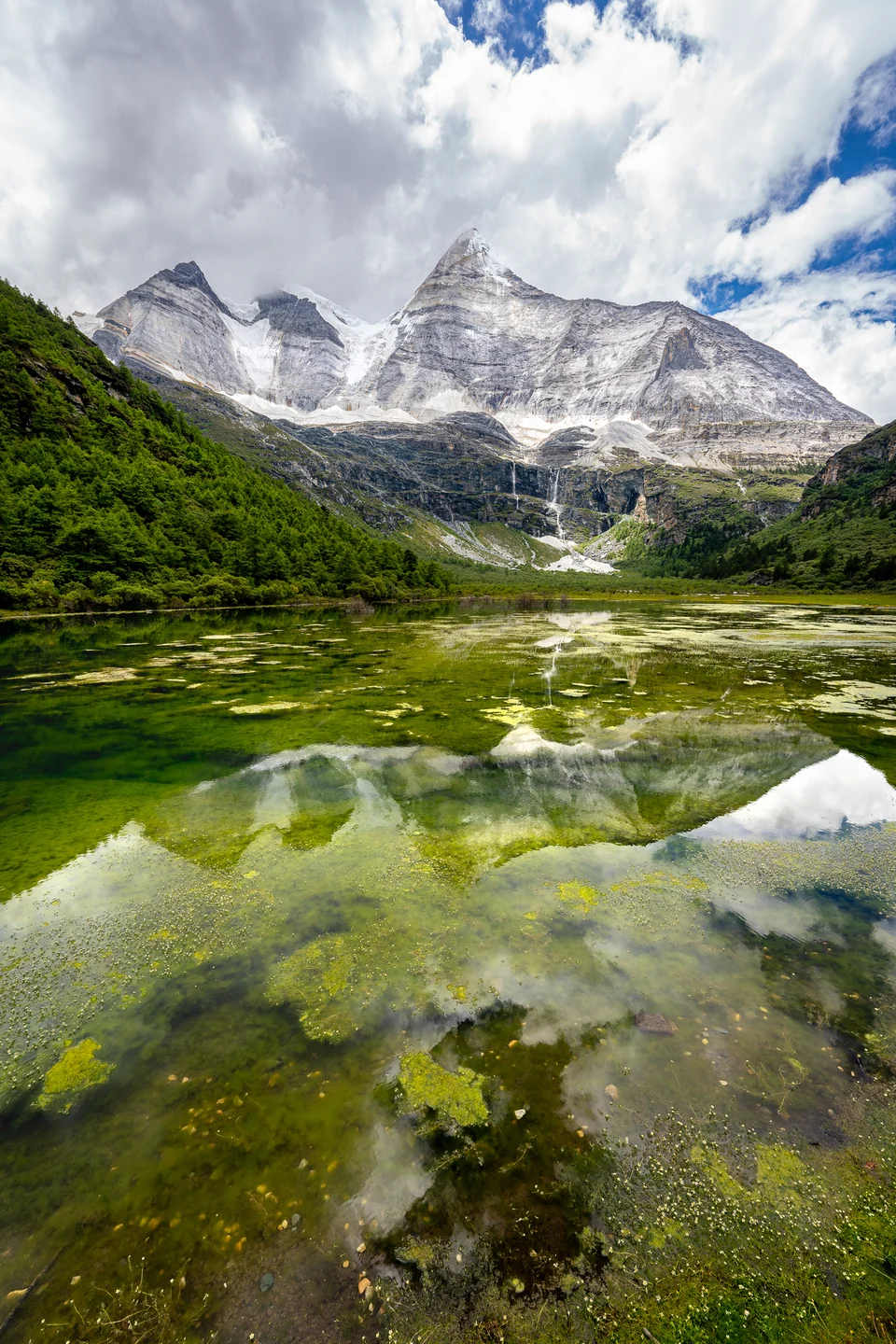 A snow-covered mountain and green valley reflected in still alpine water.