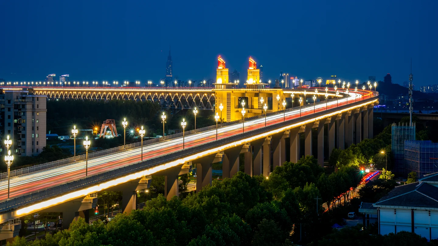 Long trails of headlights cross an illuminated bridge against a deep blue evening sky.